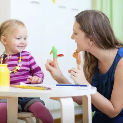 Uśmiechnięta niania bawi się z dzieckiem pacynkami na palcach. Dziewczynka ma na szyi kolorowy naszyjnik, a obok stoi żółty wazon z kredkami.