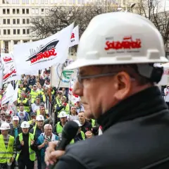 Mężczyzna w białym kasku z logo "Solidarność" przemawia do tłumu ludzi z flagami. To demonstracja związków zawodowych.