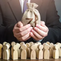 Biznesmen w garniturze trzyma worek z pieniędzmi dla grupy drewnianych ludzików. To symbol, jak siła wyższa może wpływać na losy wielu.