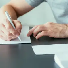 Ręka pisze długopisem na białej kartce, przygotowując się do wysłania listu motywacyjnego.