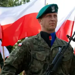 Dumnie stoi dowódca w zielonym beretcie, z karabinem i polską flagą w tle.