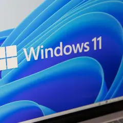 Ekran laptopa z logo Windows 11 i niebieskim, falistym tłem. System wymaga modułu TPM.