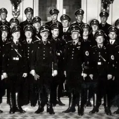Grupa mężczyzn w czarnych mundurach i czapkach z orłem, przypominających członków gestapo, pozuje do zdjęcia.
