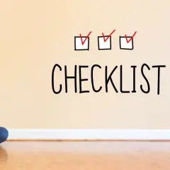 Młoda kobieta z laptopem siedzi na podłodze. Na ścianie nad nią widnieje napis "CHECKLIST" z trzema zaznaczonymi polami.