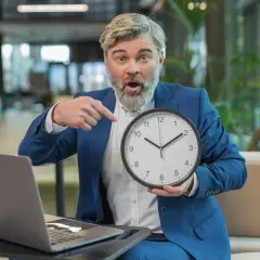 Zaskoczony mężczyzna w garniturze wskazuje na zegar, przypominając o zbliżającym się terminie.