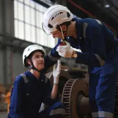 Dwóch pracowników w kombinezonach i kaskach omawia zadanie. Jeden z nich, logistyk, używa krótkofalówki.