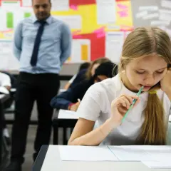Młoda abiturientka zamyślona nad arkuszem egzaminacyjnym, podczas gdy nauczyciel obserwuje klasę.