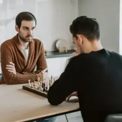 Dwóch mężczyzn gra w szachy. Jeden z nich, w brązowym swetrze, zamyślony patrzy na planszę, gotowy na gambit.