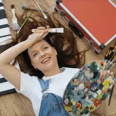 Uśmiechnięta artystka z paletą i pędzlami, otoczona płótnami i walizką. Jej pasje malarskie są widoczne w każdym detalu.