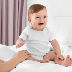 Uśmiechnięte dziecko w białym body, kiedy dziecko siada na łóżku, a dłoń rodzica delikatnie go podtrzymuje.