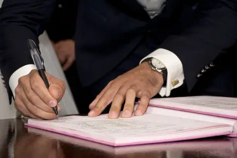 Mężczyzna w garniturze składa parafkę na dokumencie. Elegancki zegarek i spinki do mankietów podkreślają uroczystość chwili.