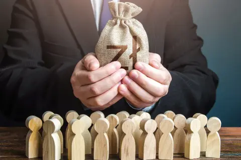 Biznesmen w garniturze trzyma worek z pieniędzmi dla grupy drewnianych ludzików. To symbol, jak siła wyższa może wpływać na losy wielu.