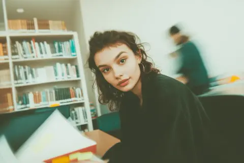 Młoda kobieta w okularach, otoczona książkami, studiuje w bibliotece. Ibiden, nauka i wiedza.