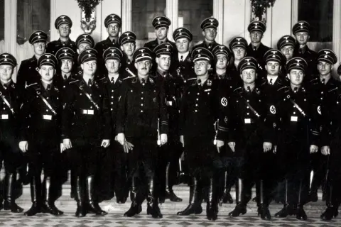 Grupa mężczyzn w czarnych mundurach i czapkach z orłem, przypominających członków gestapo, pozuje do zdjęcia.