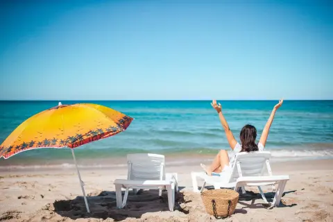 Kobieta cieszy się słońcem na plaży, zastanawiając się, ile dni urlopu jeszcze jej zostało. Obok parasol i leżaki.