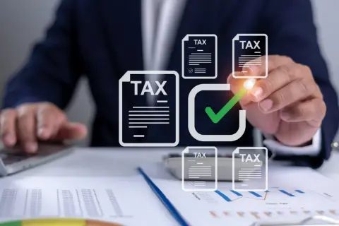 Dłoń zaznacza zielonym znacznikiem dokumenty z napisem "TAX" na tle wykresów i laptopa.