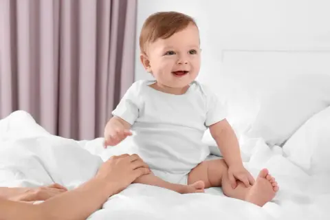Uśmiechnięte dziecko w białym body, kiedy dziecko siada na łóżku, a dłoń rodzica delikatnie go podtrzymuje.