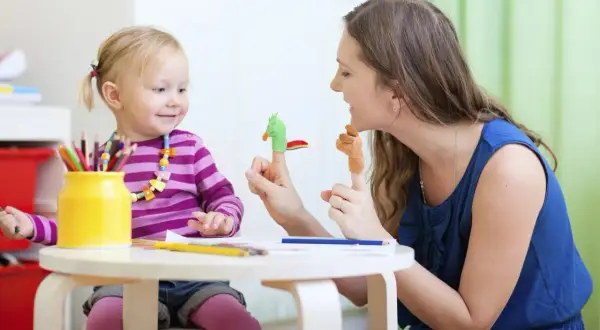 Uśmiechnięta niania bawi się z dzieckiem pacynkami na palcach. Dziewczynka ma na szyi kolorowy naszyjnik, a obok stoi żółty wazon z kredkami.