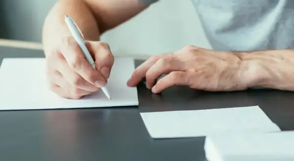 Ręka pisze długopisem na białej kartce, przygotowując się do wysłania listu motywacyjnego.