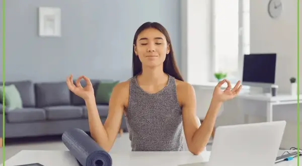 Kobieta medytuje przy biurku, osiągając work life balance. Obok leży mata do jogi i laptop.