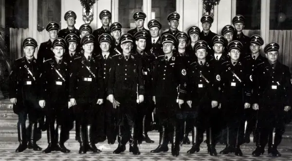 Grupa mężczyzn w czarnych mundurach i czapkach z orłem, przypominających członków gestapo, pozuje do zdjęcia.