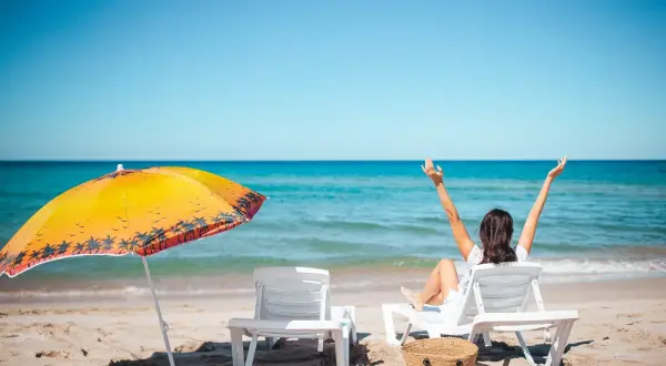 Kobieta cieszy się słońcem na plaży, zastanawiając się, ile dni urlopu jeszcze jej zostało. Obok parasol i leżaki.