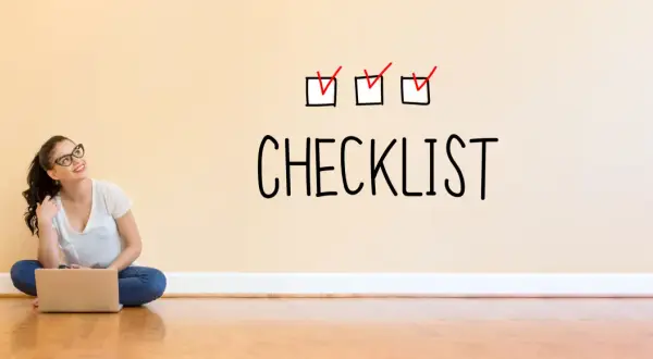 Młoda kobieta z laptopem siedzi na podłodze. Na ścianie nad nią widnieje napis "CHECKLIST" z trzema zaznaczonymi polami.