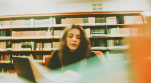 Młoda kobieta pochylona nad laptopem w bibliotece, otoczona regałami pełnymi książek. Tworzy swoją bibliografię.