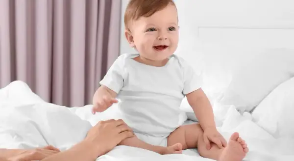 Uśmiechnięte dziecko w białym body, kiedy dziecko siada na łóżku, a dłoń rodzica delikatnie go podtrzymuje.