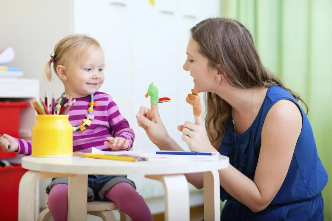 Uśmiechnięta niania bawi się z dzieckiem pacynkami na palcach. Dziewczynka ma na szyi kolorowy naszyjnik, a obok stoi żółty wazon z kredkami.