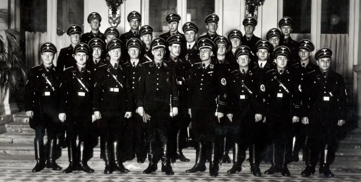 Grupa mężczyzn w czarnych mundurach i czapkach z orłem, przypominających członków gestapo, pozuje do zdjęcia.
