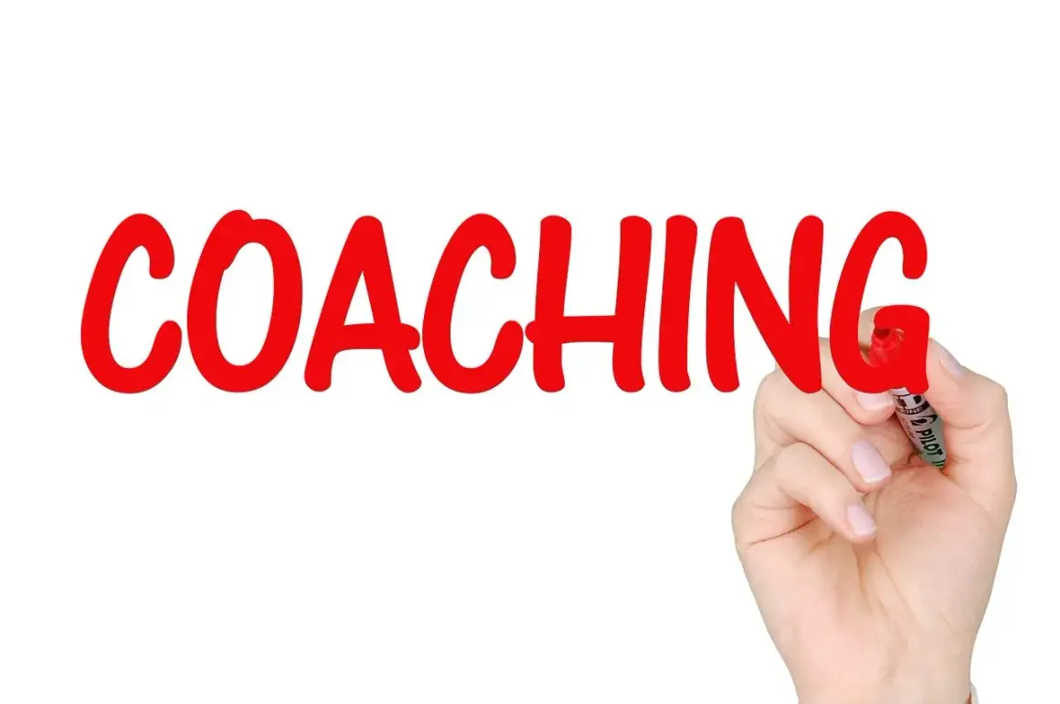 Ręka pisze czerwonym markerem słowo "COACHING". To proces rozwoju, który może obejmować lingwistykę.