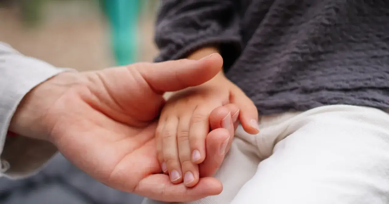 Dłoń dorosłego obejmuje małą, dziecięcą dłoń. To czuły gest, który przypomina o niewinności i infantylnym świecie dziecka.