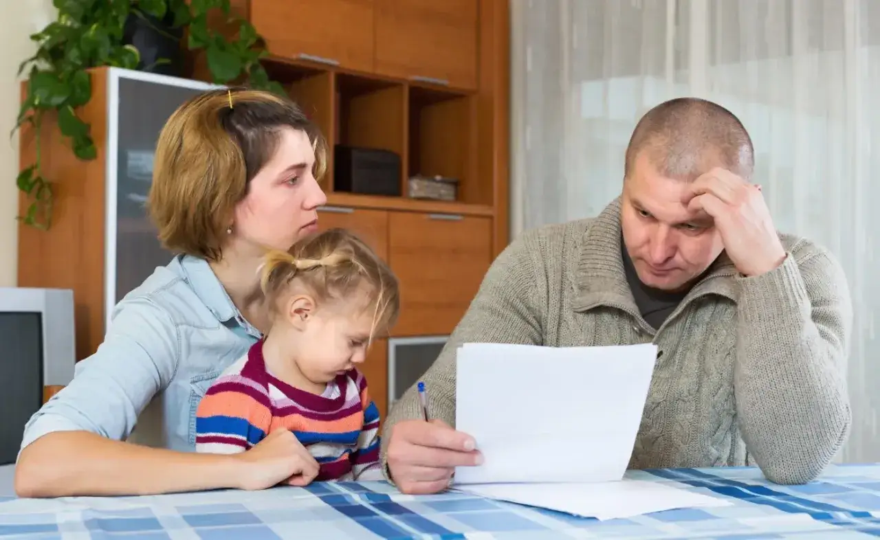 Rodzina zmartwiona rachunkami. Ojciec przegląda dokumenty, matka z córką obok. Może potrzebna będzie niania, by zaoszczędzić?