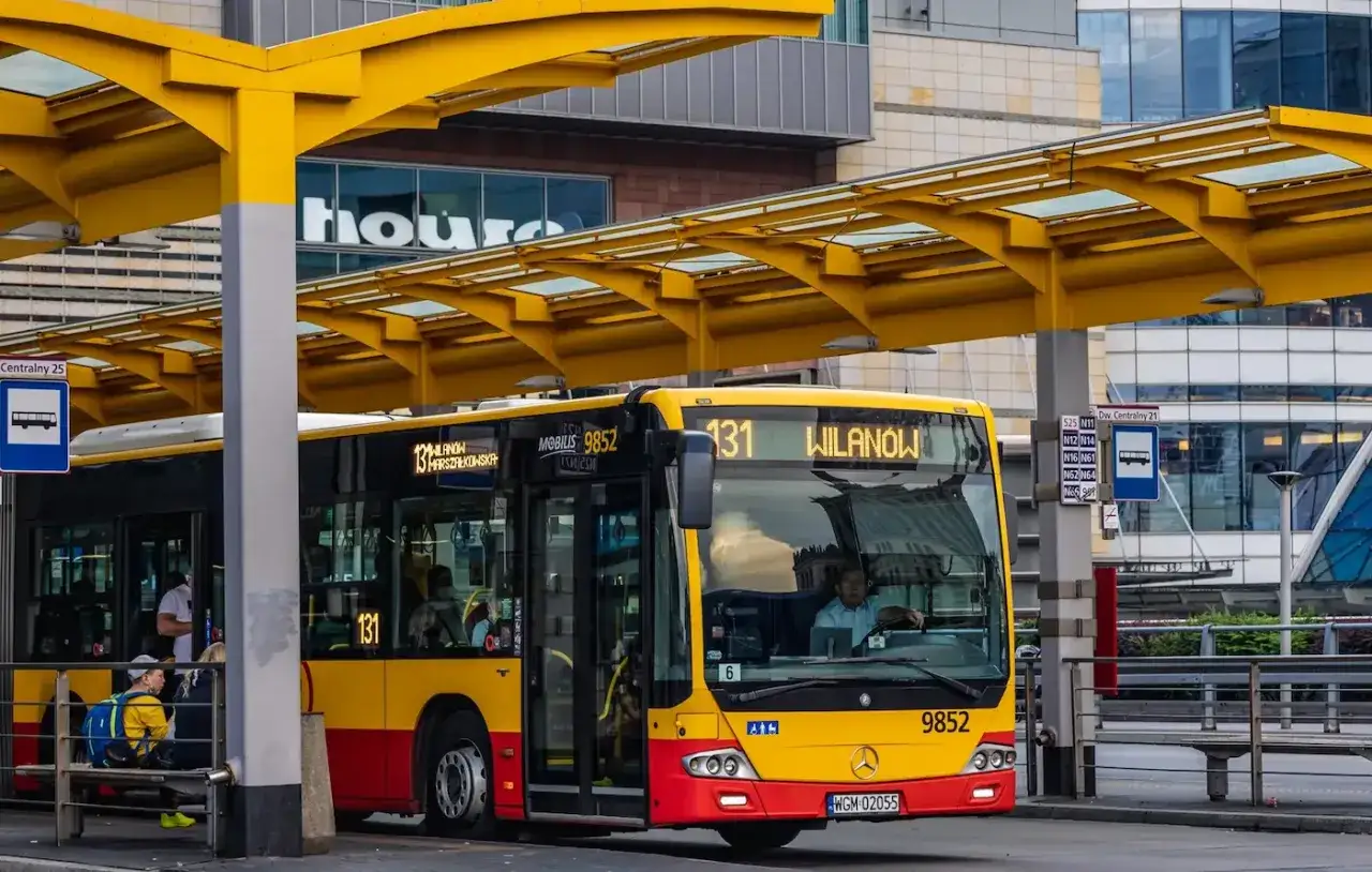 Żółto-czerwony autobus miejski na przystanku. Kierowca autobusu, jak i pasażerowie, zastanawiają się, ile zarabia kierowca autobusu.