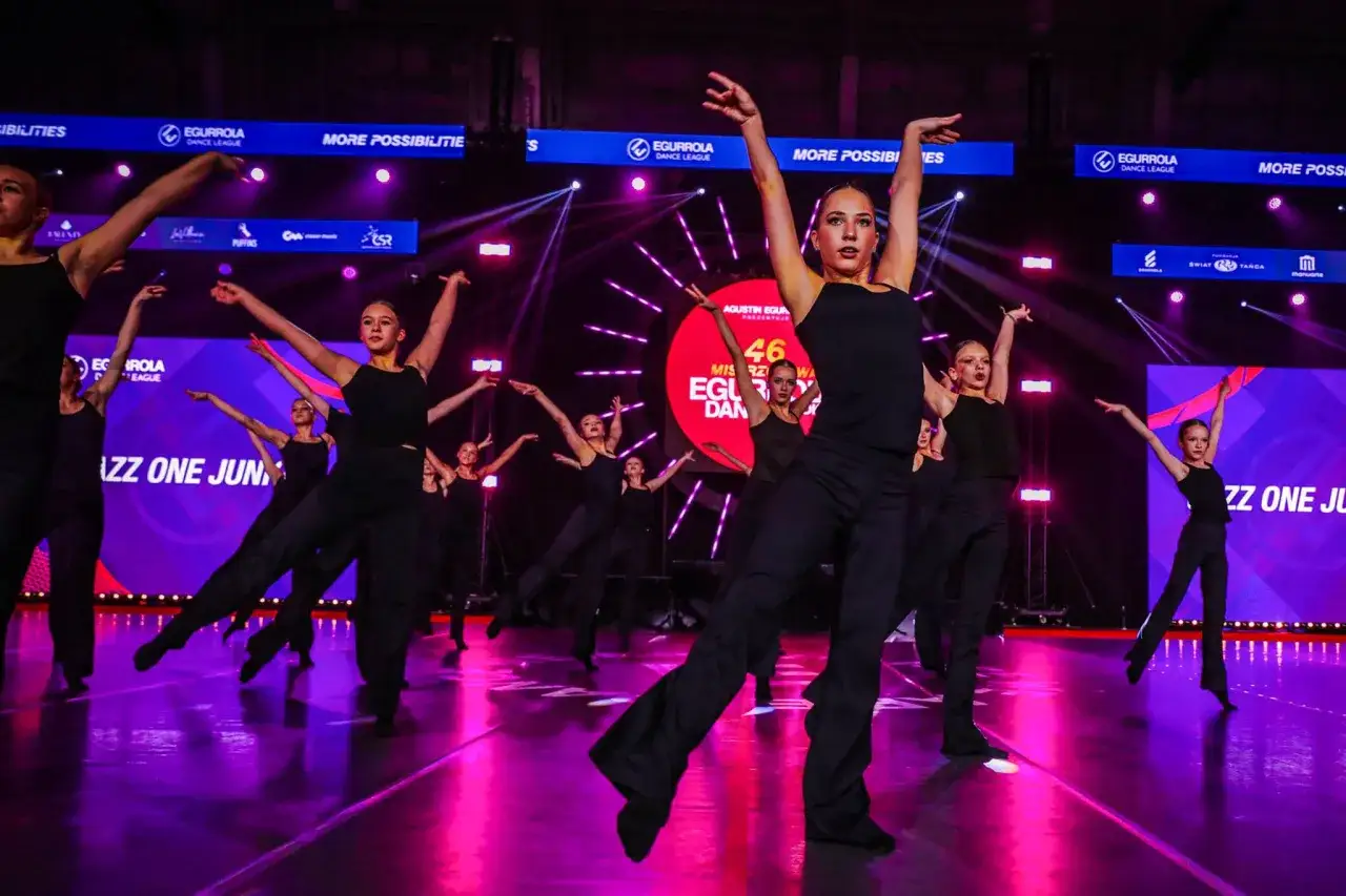 Młodzi tancerze w czarnych strojach wykonują choreografię na scenie oświetlonej fioletowym światłem.