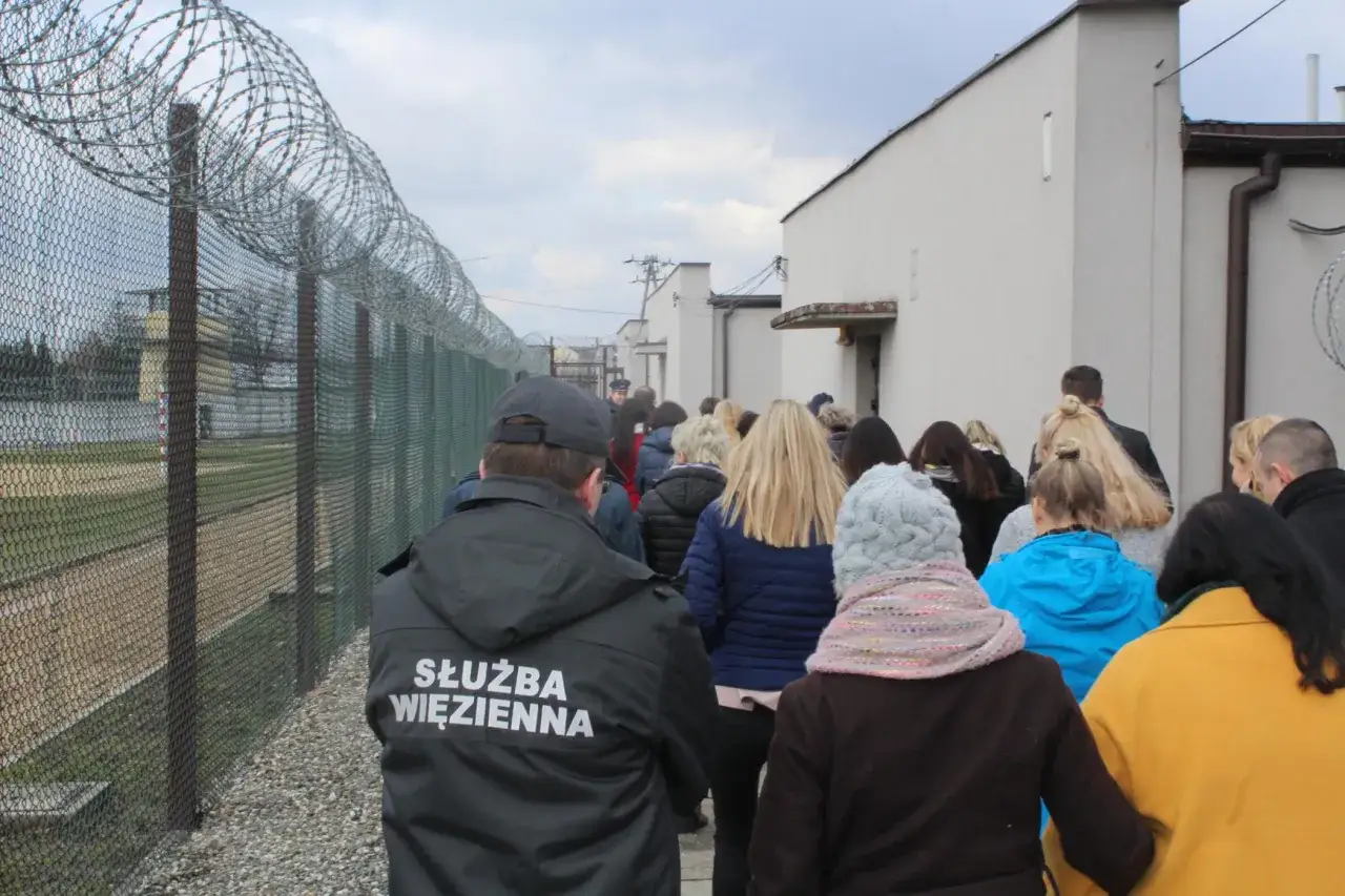 Grupa osób idzie wzdłuż ogrodzenia z drutem kolczastym. Strażnik więzienny w kurtce z napisem