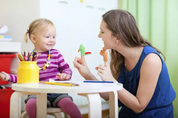 Uśmiechnięta niania bawi się z dzieckiem pacynkami na palcach. Dziewczynka ma na szyi kolorowy naszyjnik, a obok stoi żółty wazon z kredkami.