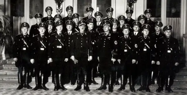 Grupa mężczyzn w czarnych mundurach i czapkach z orłem, przypominających członków gestapo, pozuje do zdjęcia.