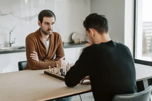 Dwóch mężczyzn gra w szachy. Jeden z nich, w brązowym swetrze, zamyślony patrzy na planszę, gotowy na gambit.