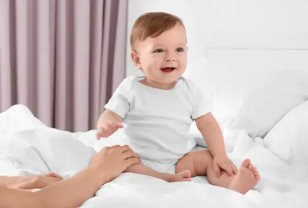 Uśmiechnięte dziecko w białym body, kiedy dziecko siada na łóżku, a dłoń rodzica delikatnie go podtrzymuje.